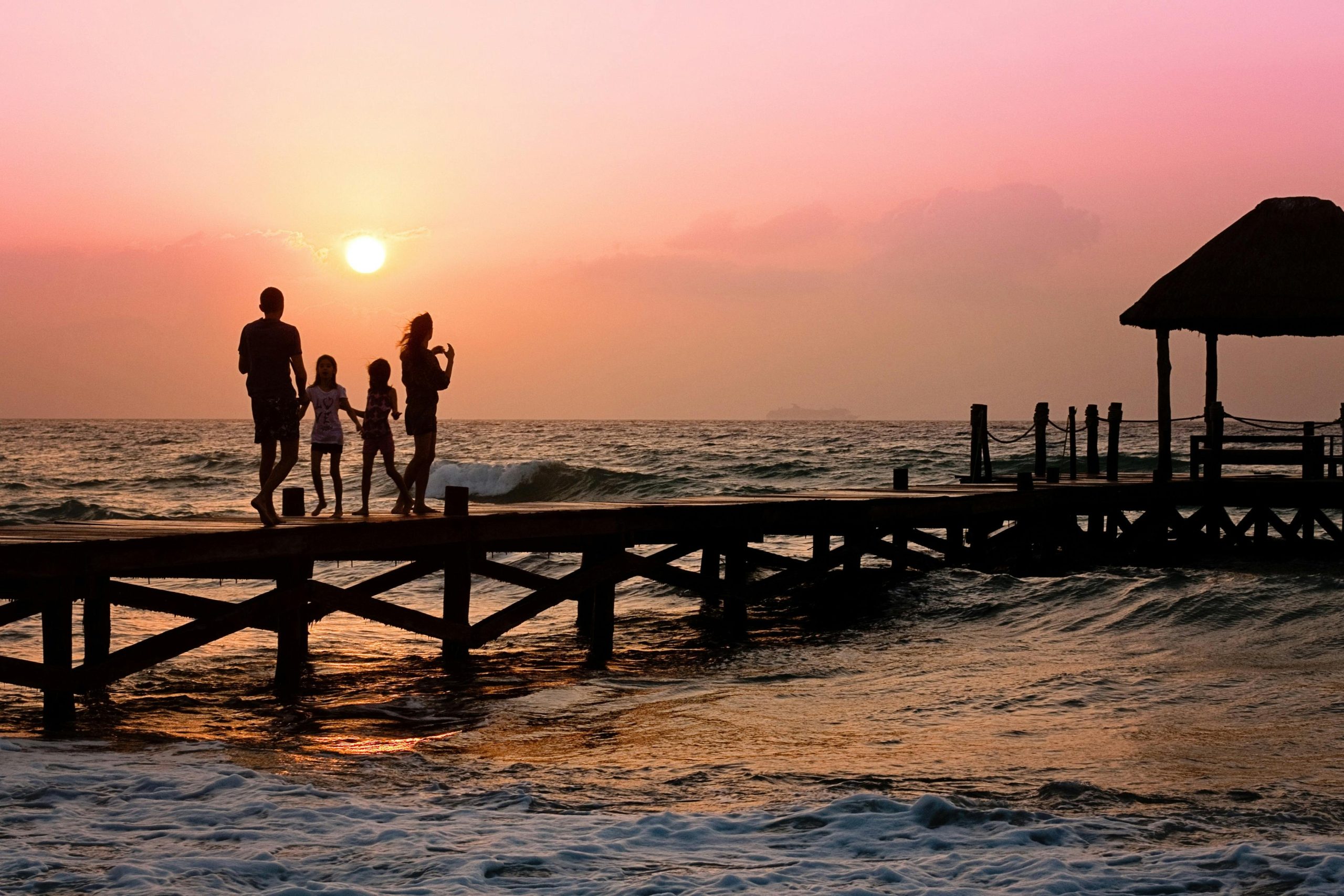 Home Silhouetted family enjoys a stroll on the beach pier at a vibrant sunset over the ocean waves.